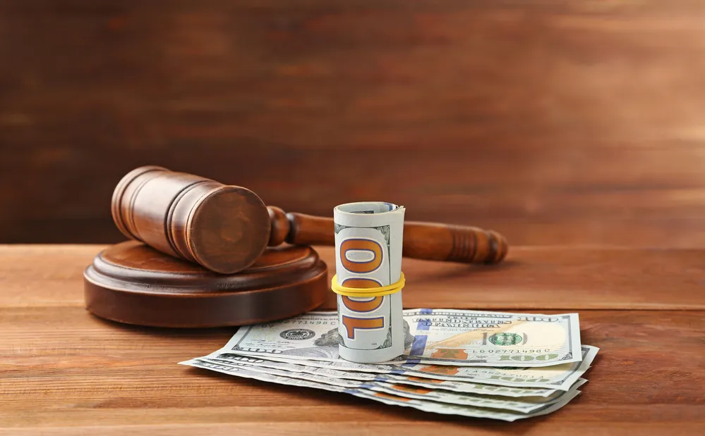 A gavel next to a roll of money and dollar bills on a wooden surface